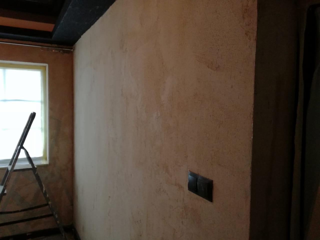 Wnętrze pokoju w trakcie prac wykończeniowych, widoczna ściana z nałożoną fakturą, drabina przy oknie i czarny włącznik światła.