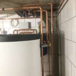 Nowoczesna kotłownia z miedzianymi rurami, białym bojlerem i szarą rurą kanalizacyjną, widok na surowy betonowy sufit i białe płytki ścienne.