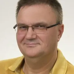 Portret mężczyzny w okularach i żółtej koszulce polo, uśmiechającego się do kamery na jasnym tle.