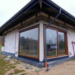 FPUH SALAMANDRA s.c. - Surowy stan domu z brązowymi oknami PCV i orynnowaniem, widok z zewnątrz na elewację z pustaków, teren budowy z narzędziami i resztkami materiałów.