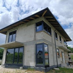 FPUH SALAMANDRA s.c. - Surowy budynek z szarego betonu komórkowego z zamontowanymi oknami w grafitowych ramach, widok z zewnątrz w słoneczny dzień.