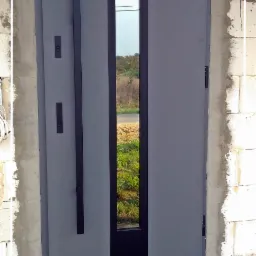 Nowoczesne, szare drzwi wejściowe z długim, pionowym uchwytem i wąskim przeszkleniem, osadzone w surowym otworze murarskim, z widoczną warstwą izolacji i logo producenta w rogu.