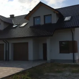 Nowoczesny dom jednorodzinny z dwustanowiskowym garażem, ciemną dachówką i stolarką okienną w odcieniach brązu, widziany z poziomu chodnika.