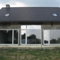 FPUH SALAMANDRA s.c. - Dom w trakcie budowy z zamontowanymi białymi oknami, widoczne rusztowanie wewnątrz, szara dachówka i okna dachowe.