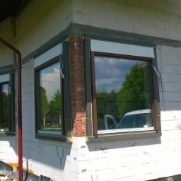 FPUH SALAMANDRA s.c. - Surowy stan elewacji domu z zainstalowanymi oknami z brązowymi ramami i roletami zewnętrznymi, widoczne pozostałości po pracach murarskich i instalacyjnych.