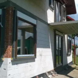 Budynek w trakcie budowy z zamontowanymi oknami z brązowymi ramami i częściowo opuszczonymi roletami zewnętrznymi, widoczne cegły i bloczki fundamentowe.