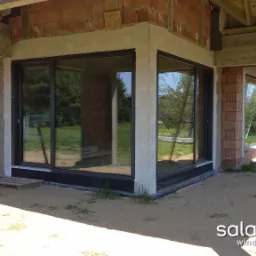 Surowy stan budynku z zamontowanymi oknami PCV z czarnymi ramami, widoczne cegły i betonowe elementy konstrukcyjne, ziemia na zewnątrz, logo Salamandra windows & doors w prawym dolnym rogu.