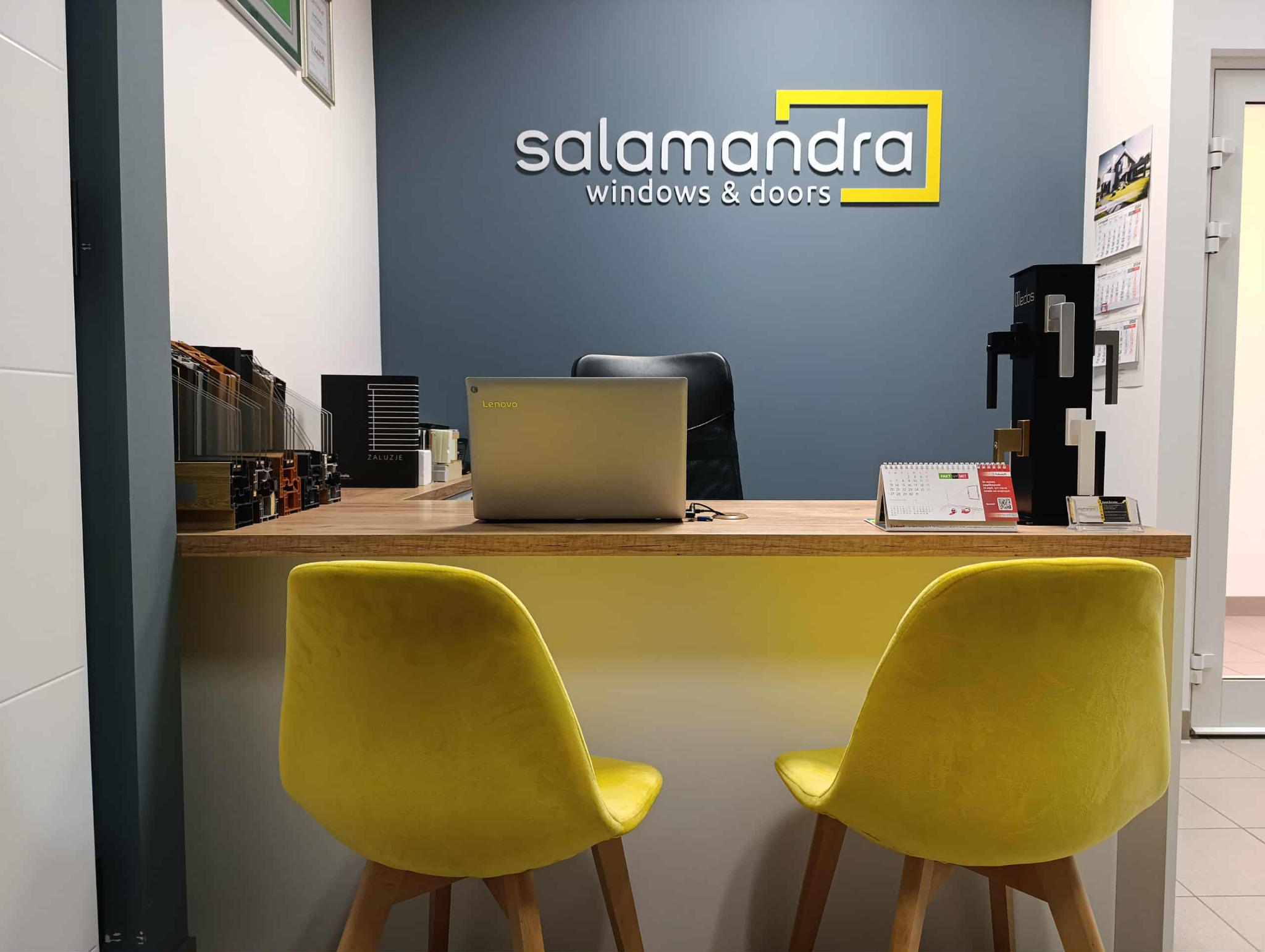 Biuro firmy Salamandra Windows & Doors z ekspozycją profili okiennych, próbek klamek i laptopem Lenovo na drewnianym biurku. Żółte krzesła dla klientów.