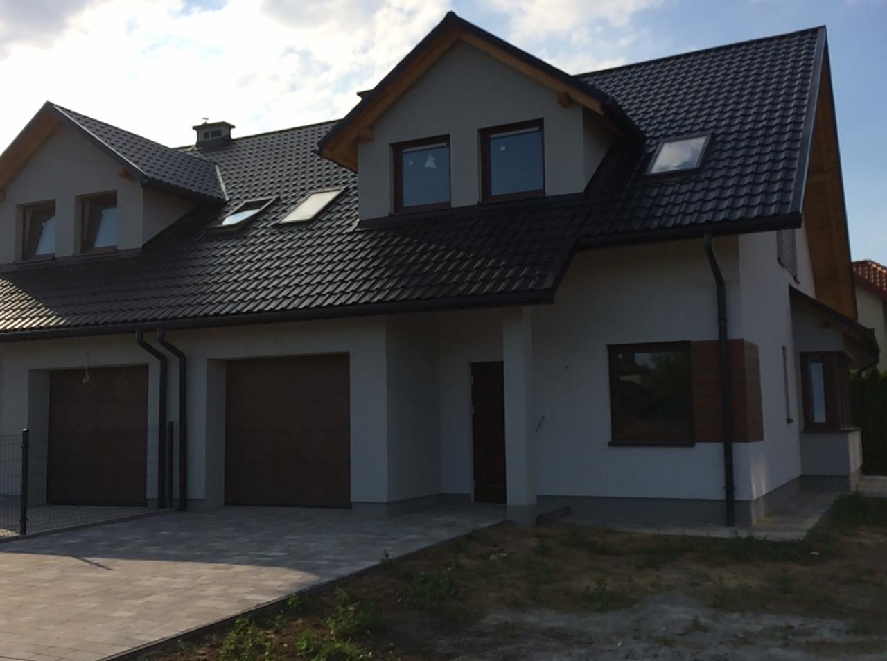 Nowoczesny dom jednorodzinny z dwustanowiskowym garażem, ciemną dachówką i stolarką okienną w odcieniach brązu, widziany z poziomu chodnika.