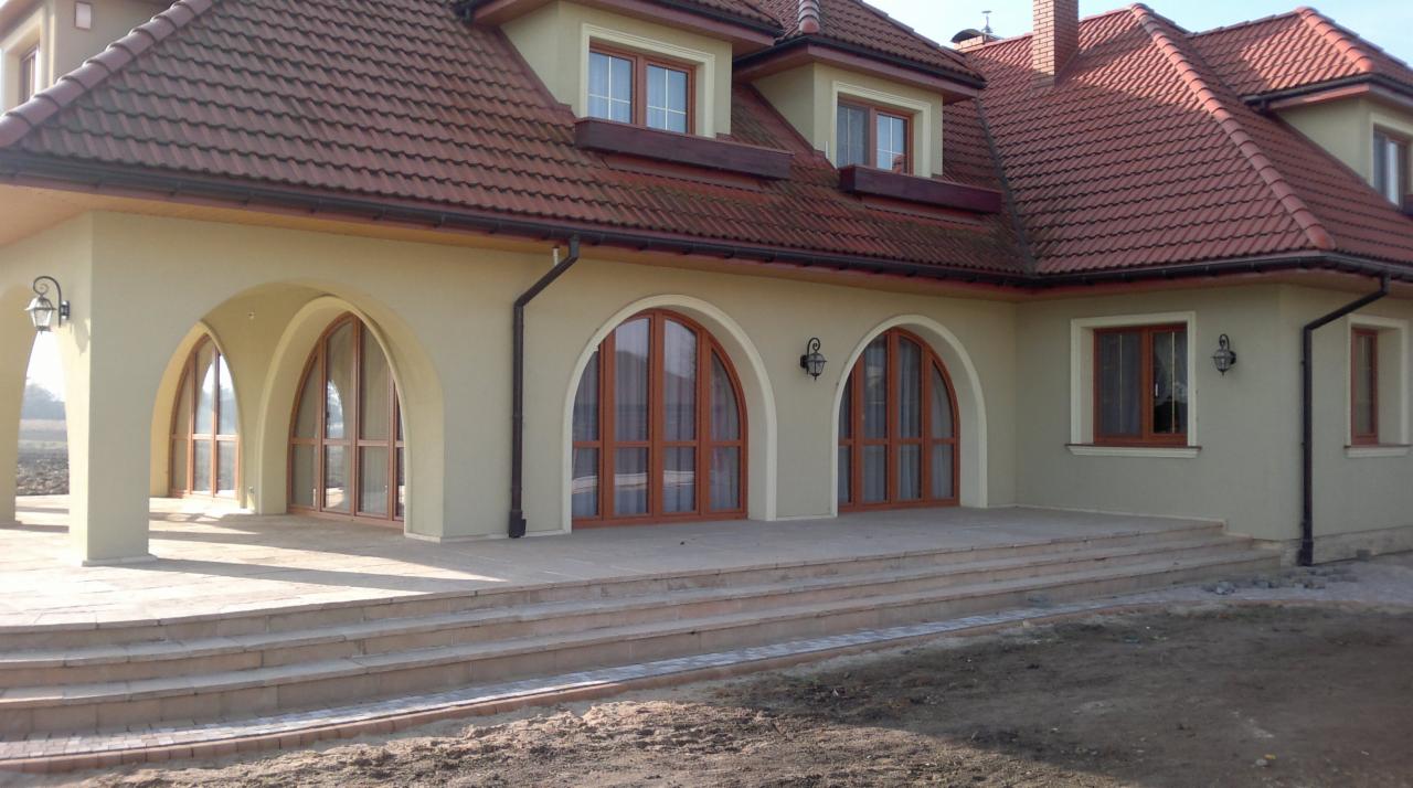 Elewacja domu z łukowymi oknami i drzwiami balkonowymi w drewnianej ramie, widok z zewnątrz na taras wyłożony płytkami.