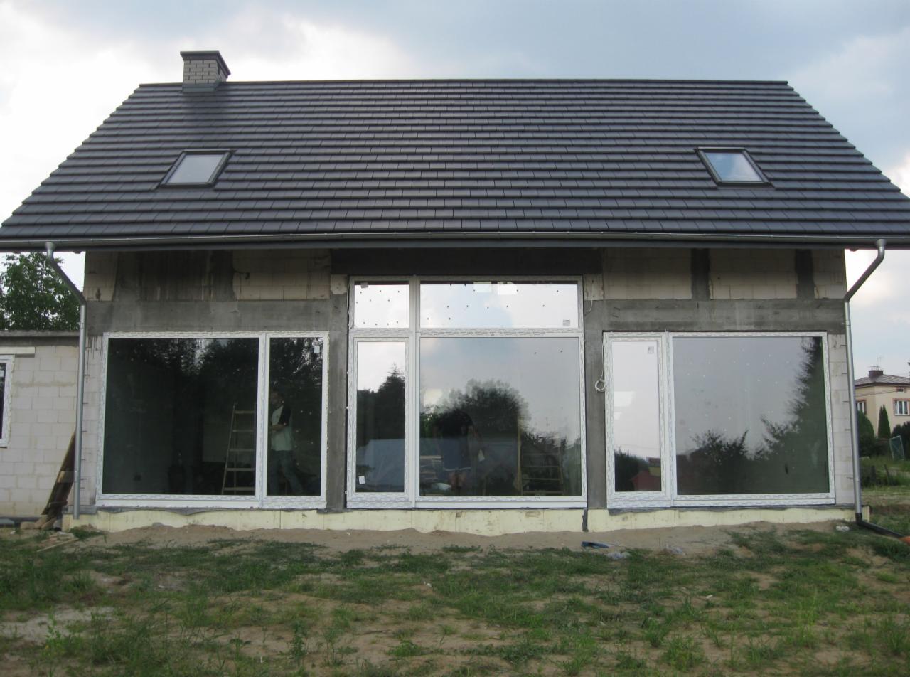 Dom w trakcie budowy z zamontowanymi białymi oknami, widoczne rusztowanie wewnątrz, szara dachówka i okna dachowe.