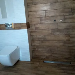 Nowoczesna toaleta podwieszana w łazience wykończonej płytkami imitującymi drewno, z widocznym odpływem liniowym i chromowanym przyciskiem spłukującym.