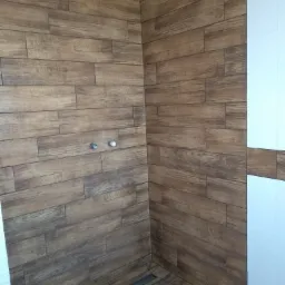 Kąt w kabinie prysznicowej wyłożony płytkami imitującymi drewno, z widocznym odpływem liniowym w podłodze i fragmentem białych płytek na ścianie.