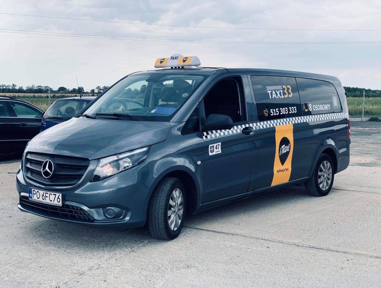 Szary van taxi z numerem 33, numerem telefonu i logo aplikacji na boku, zaparkowany na betonowym placu w pochmurny dzień. Widoczny numer rejestracyjny.