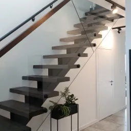 Nowoczesne, wiszące stopnie w odcieniach szarości, zabezpieczone szklaną balustradą z czarną poręczą, uzupełnione rośliną doniczkową w minimalistycznej, czarnej osłonce.