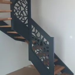 Nowoczesne schody z drewnianymi stopniami i metalową balustradą o geometrycznym wzorze, pomalowaną na ciemny kolor, widoczne z dołu, na tle białej ściany.