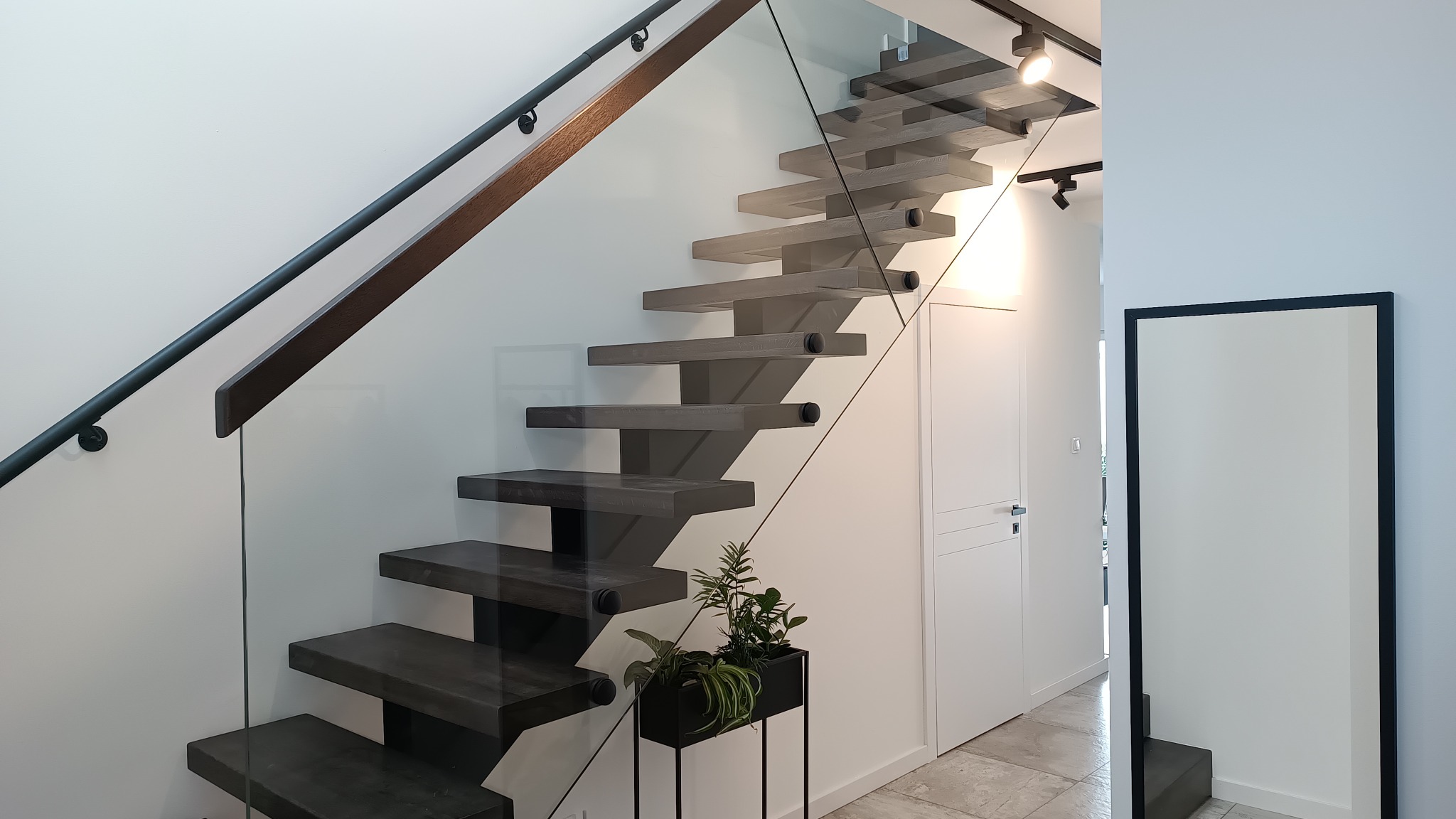 Nowoczesne, wiszące stopnie w odcieniach szarości, zabezpieczone szklaną balustradą z czarną poręczą, uzupełnione rośliną doniczkową w minimalistycznej, czarnej osłonce.