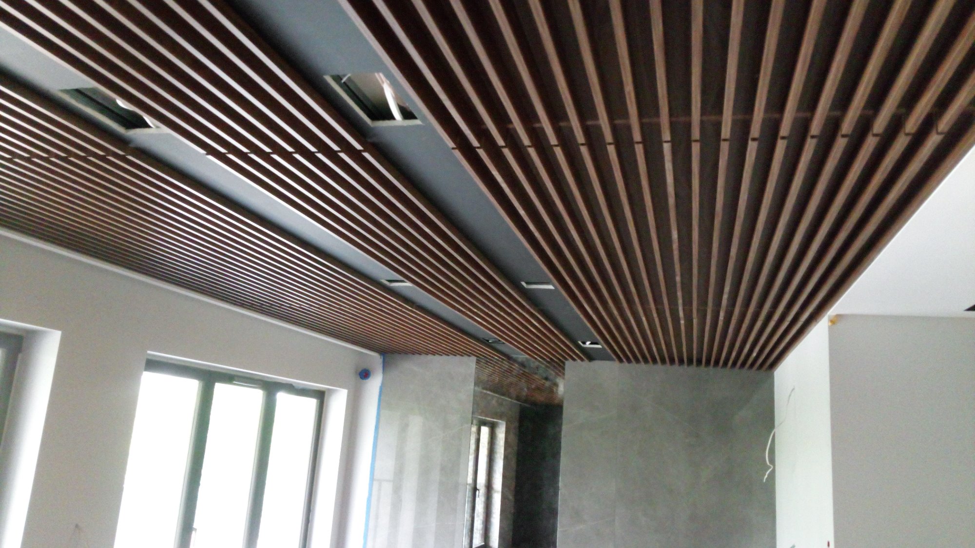 Drewniane lamele sufitowe w nowoczesnym wnętrzu, widoczne okna i szara ściana.