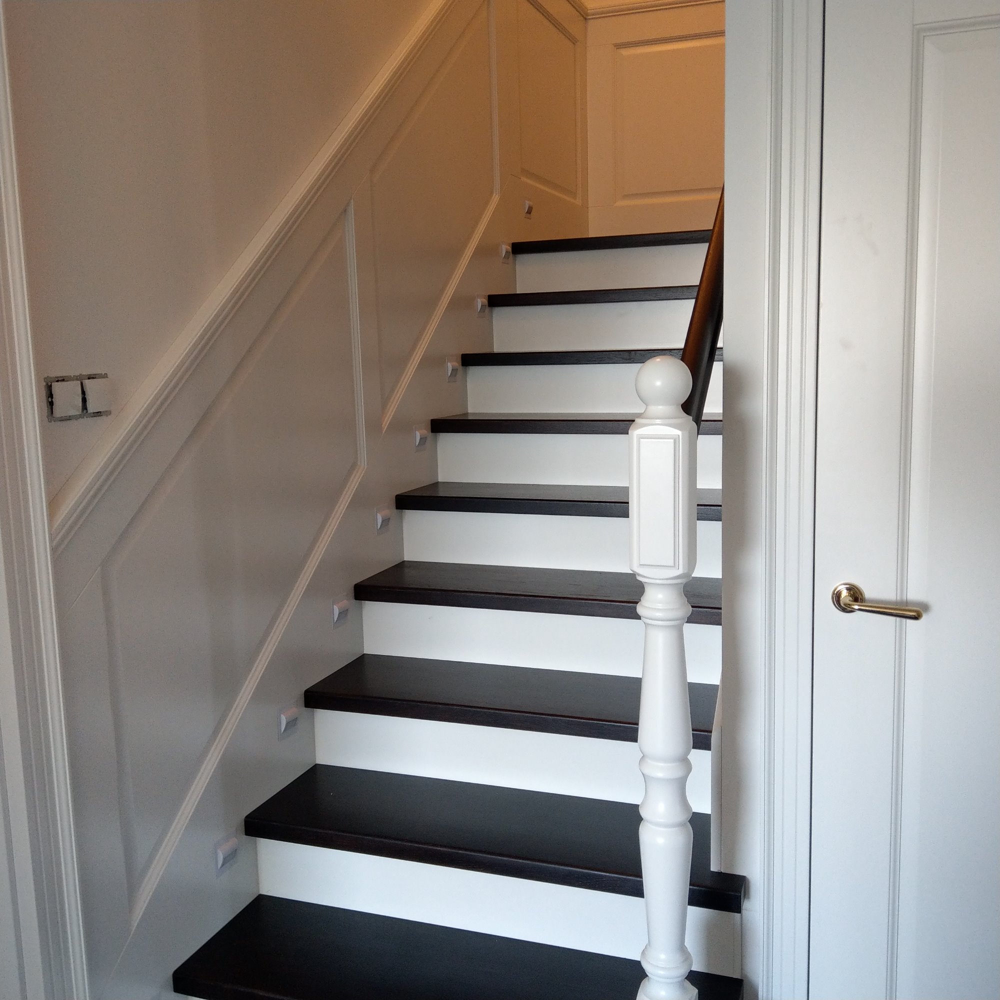 Klasyczne schody wewnętrzne z ciemnymi stopniami i białymi podstopnicami, biała balustrada z ozdobnym tralką, ściany wykończone boazerią, widoczny fragment instalacji elektrycznej.