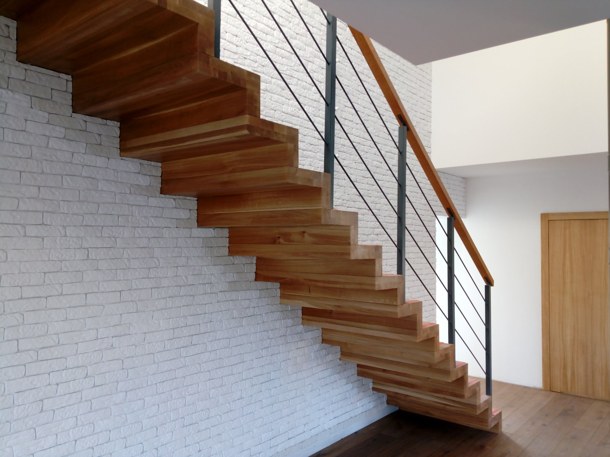 Nowoczesne, ażurowe schody z drewna, wspornikowe, z balustradą z czarnych prętów i drewnianą poręczą, na tle białej ściany z cegieł i drewnianych drzwi.