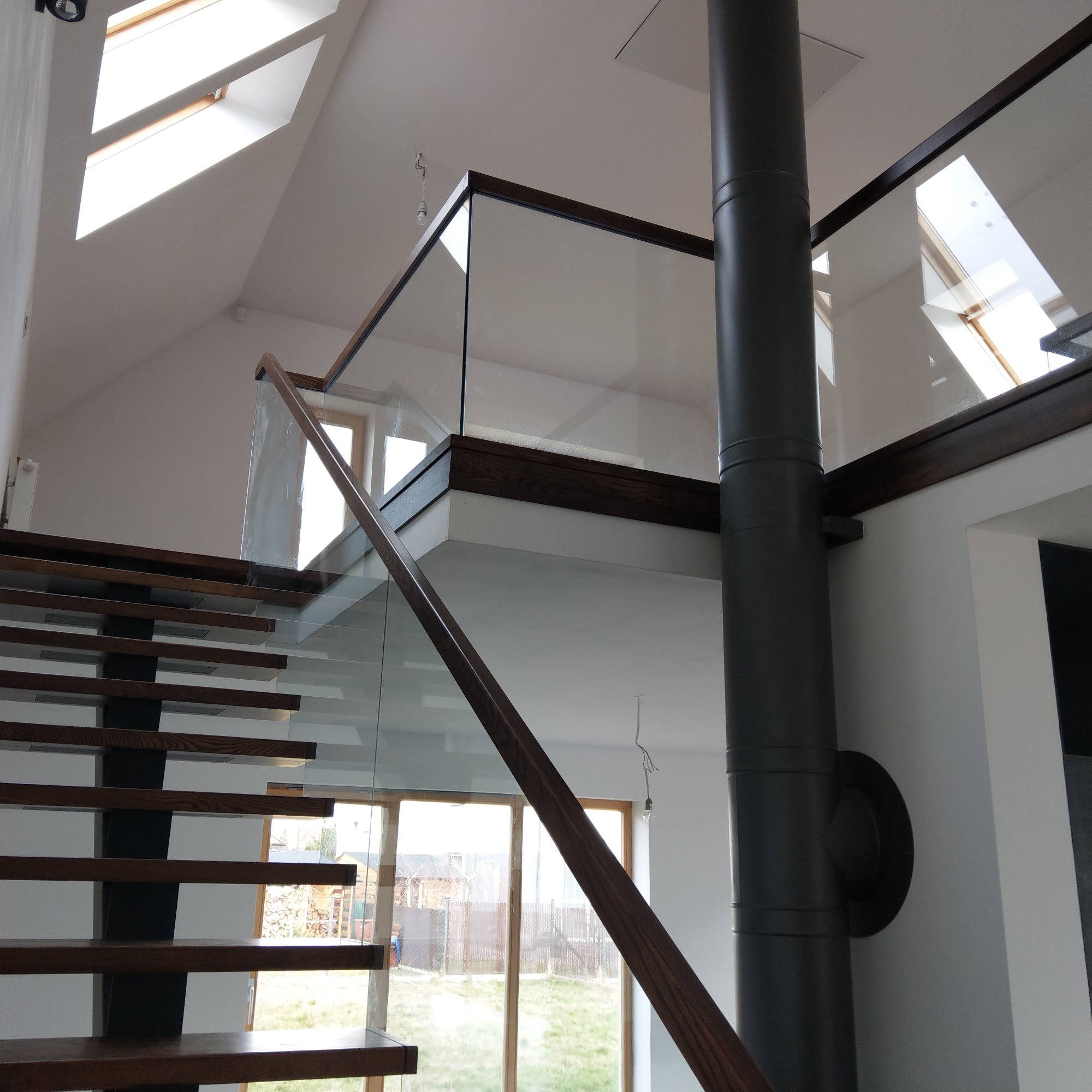 Nowoczesne schody z drewnianymi stopniami i czarnymi wspornikami, szklane balustrady z ciemnobrązowymi poręczami, widok z dołu na poddasze z oknami dachowymi i kominem.