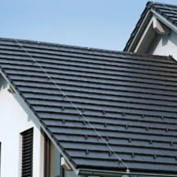 Grafitowy dach z dachówki ceramicznej na dwuspadowym domu z białą elewacją i ciemnymi oknami, widok z dołu w słoneczny dzień.