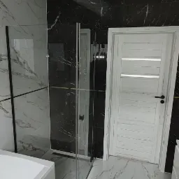 Nowoczesna łazienka z czarno-białymi marmurkowymi płytkami, szklaną kabiną prysznicową z czarnymi ramami i białą, drewnopodobną stolarką drzwiową. Widoczny fragment wanny i otwór na oświetlenie...