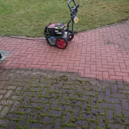 Częściowo wyczyszczony z mchu i zabrudzeń czerwony chodnik z kostki, widoczna myjka ciśnieniowa z czerwonymi kołami i wąż, fragment ściany budynku z rynną.