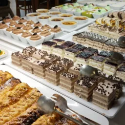 Bogaty bufet deserowy z różnorodnymi ciastami, kremami i deserami w małych miseczkach, widoczne szczypce do nakładania.