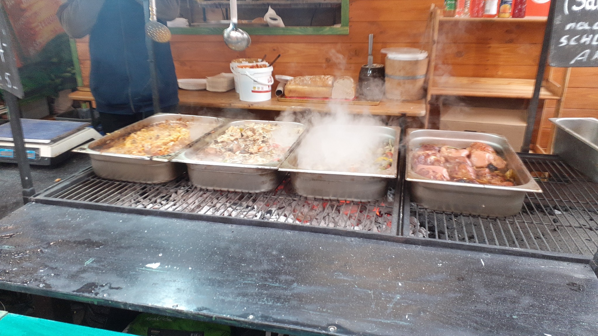 Cztery metalowe pojemniki z różnymi potrawami, w tym mięsem, podgrzewane na ruszcie nad żarzącymi się węglami, z widocznym parą i drewnianym stoiskiem w tle.