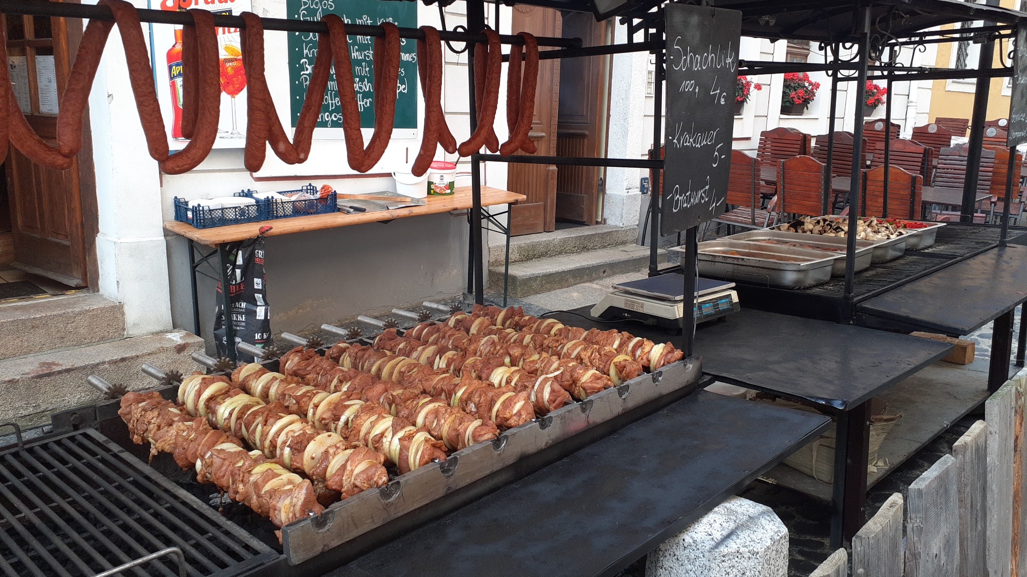 Stragan z grillem na zewnątrz: szaszłyki z mięsa i cebuli na ruszcie, wiszące wędliny, tablica z menu i ceny, metalowe pojemniki z warzywami, stoliki i krzesła w tle.