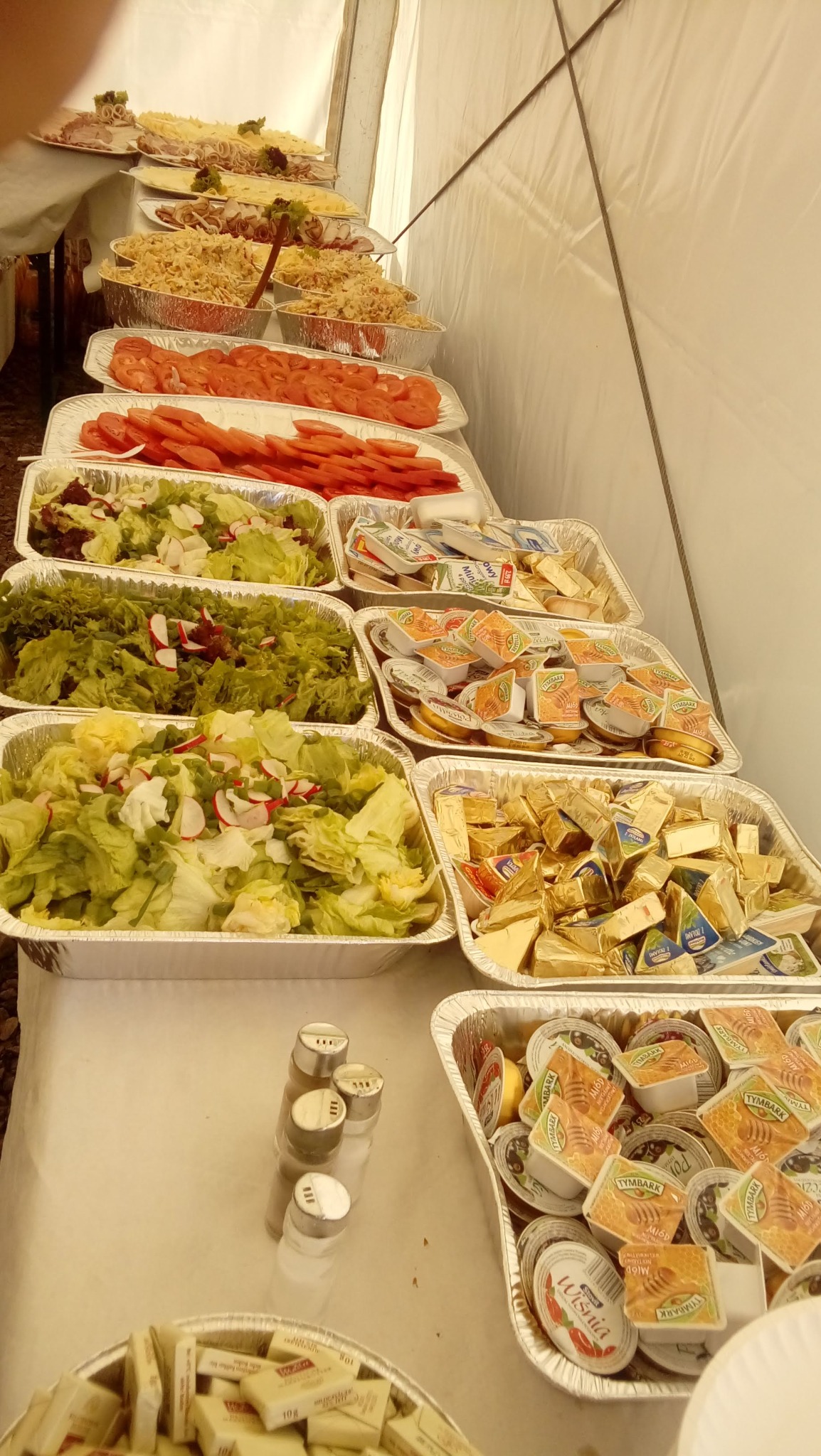 Stół cateringowy w namiocie, zastawiony aluminiowymi tackami z sałatkami, plastrami pomidorów, serami w różnych opakowaniach i wędlinami na paterach, widoczne przyprawy w solniczkach.