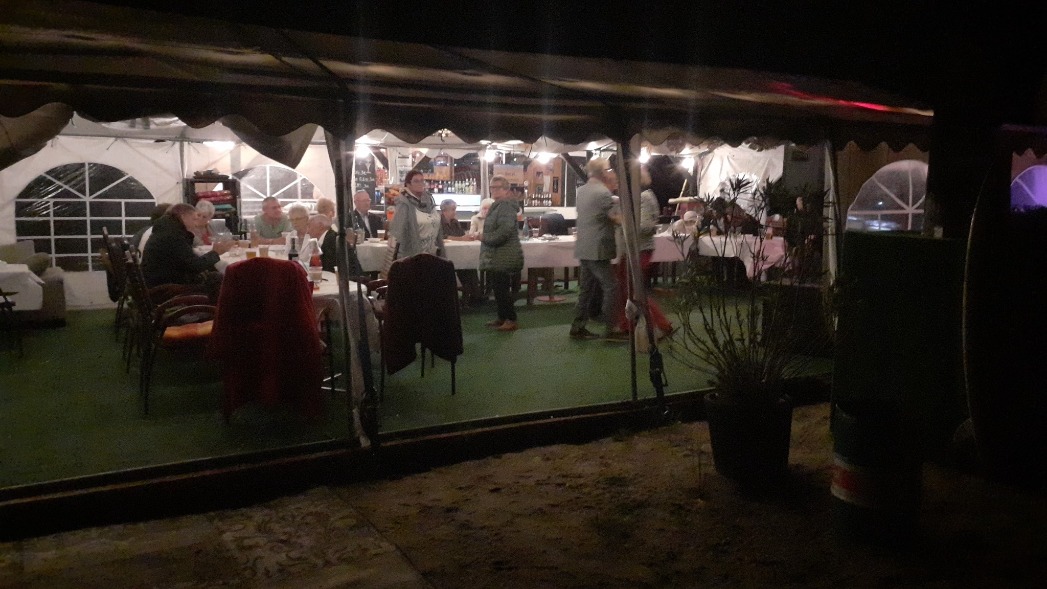 Uroczystość pod namiotem: goście siedzą przy stołach z białymi obrusami, bar z alkoholem w tle, widoczne oświetlenie i dekoracje.