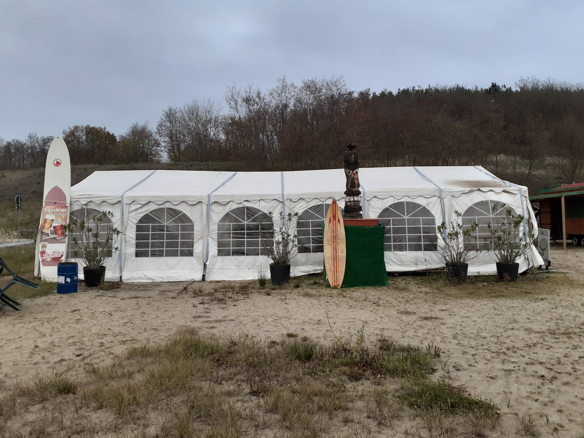 Duży biały namiot z oknami na piaszczystej plaży, dekoracje w stylu surfingowym: deski, rzeźba tiki, rośliny w donicach.