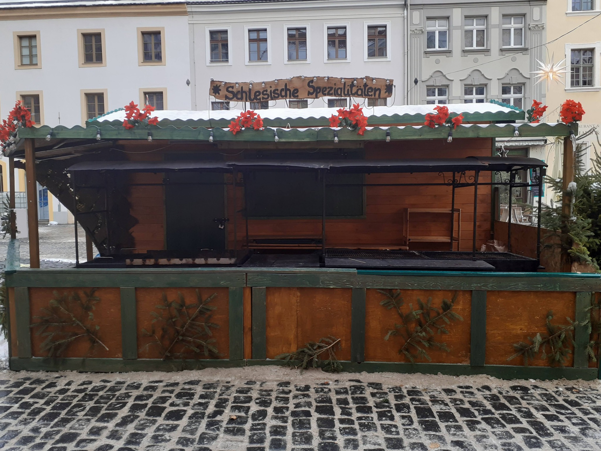 Zewnętrzny drewniany stragan z napisem 'Schlesische Spezialitäten', udekorowany czerwonymi kwiatami i gałązkami świerkowymi, stoi na brukowanym placu w zimowej scenerii.