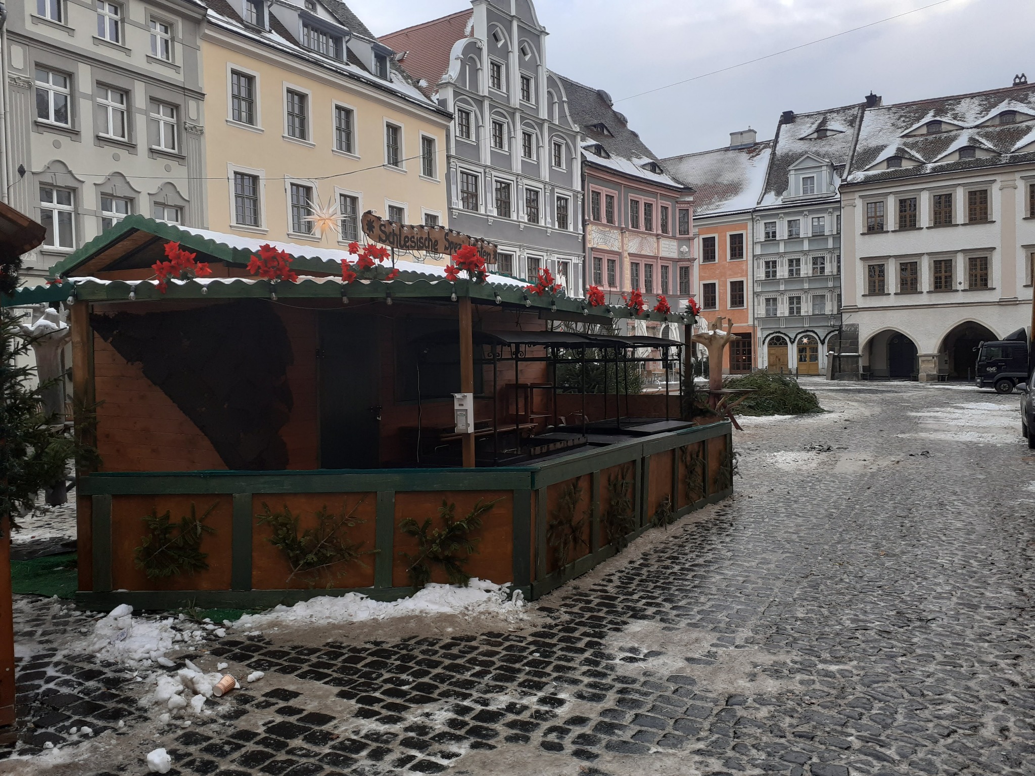 Drewniany stragan na rynku miejskim, udekorowany czerwonymi kwiatami, z widocznymi śladami śniegu na brukowanej nawierzchni i historycznymi kamienicami w tle.