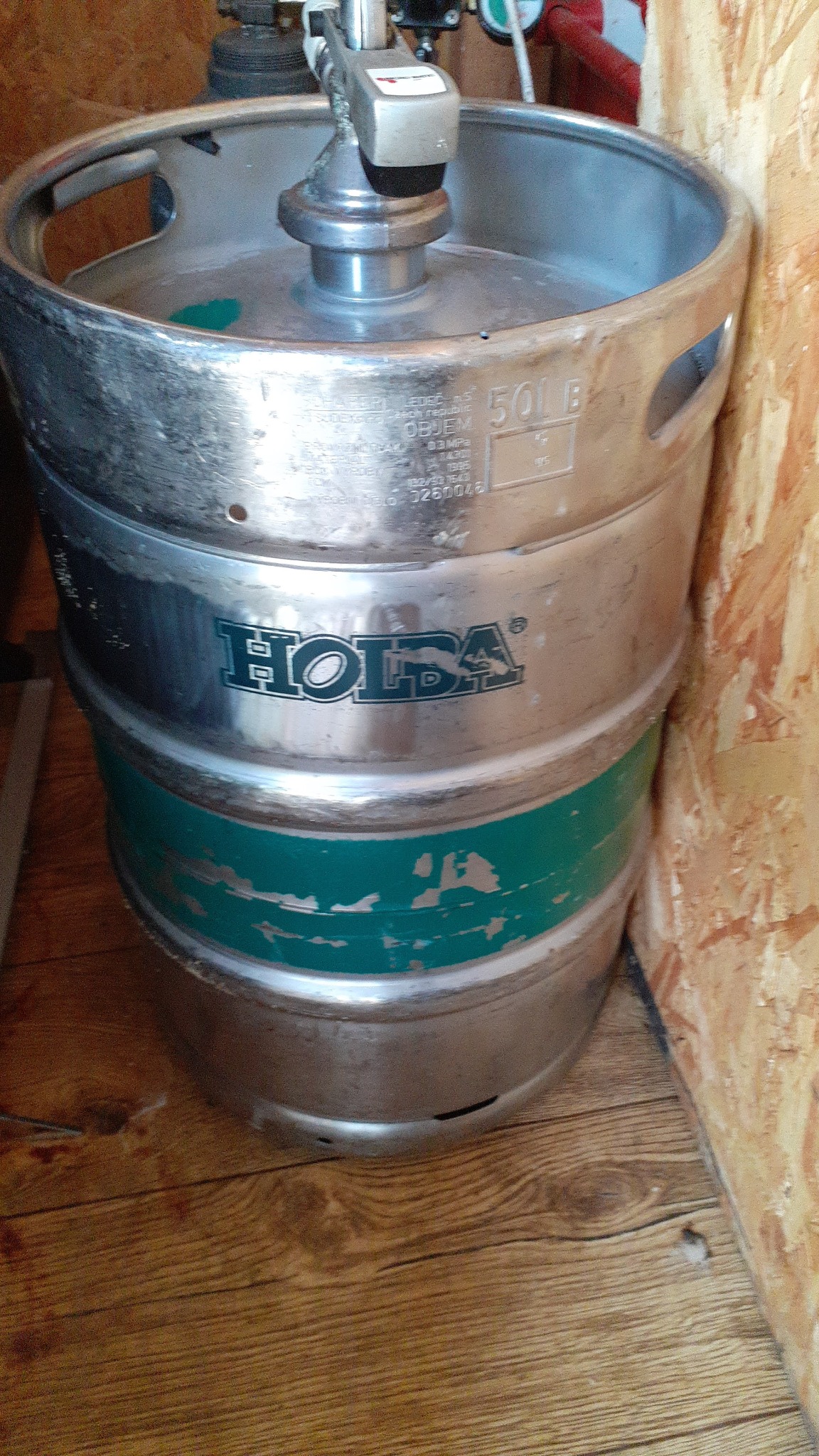 Metalowa beczka z logiem 'Holsa' i zielonymi pasami, stojąca na drewnianej podłodze przy ścianie wyłożonej płytami drewnianymi.