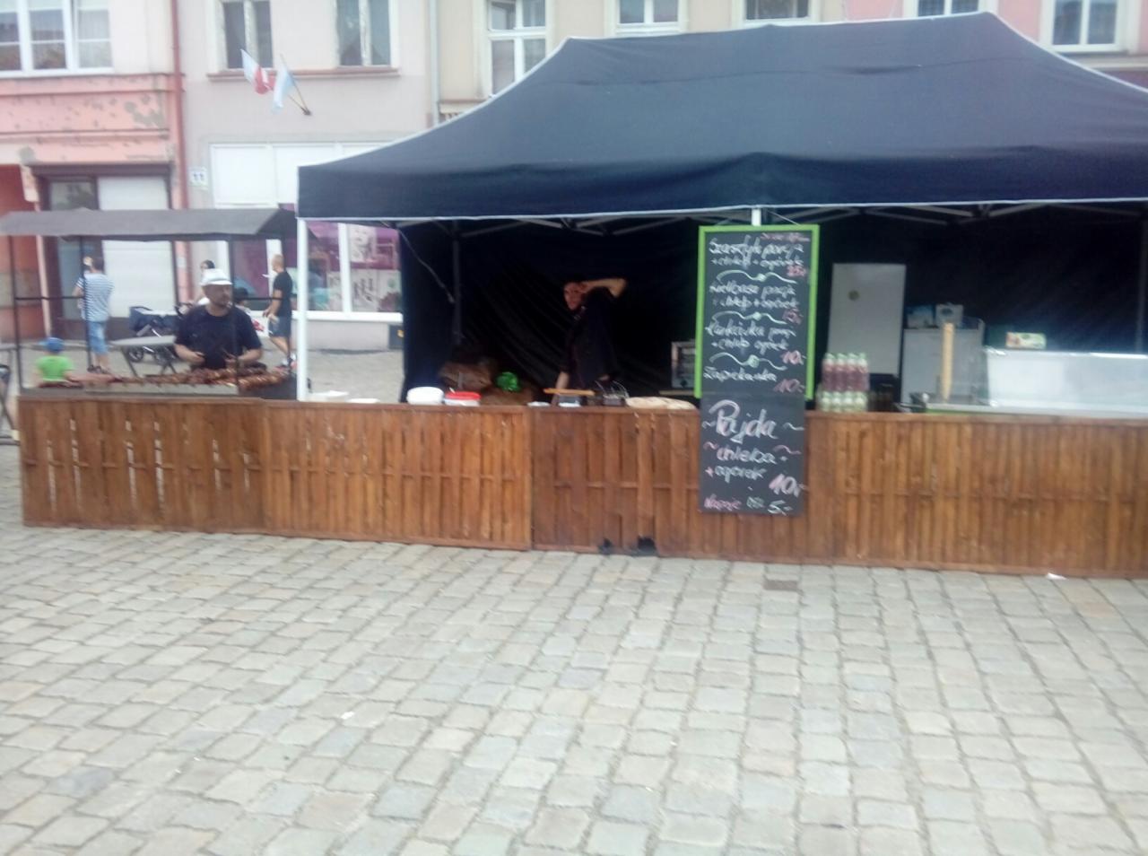 Mobilne stoisko gastronomiczne z ciemnym namiotem i drewnianym frontem, oferujące dania z grilla i zapiekanki, z menu wypisanym kredą na tablicach.
