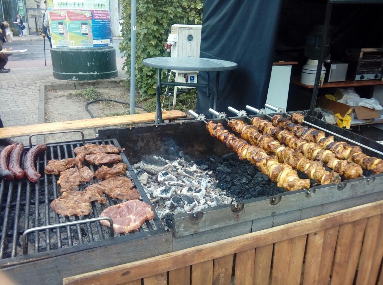 Grillowanie mięs i szaszłyków na dużym ruszcie z węglem drzewnym, widoczne kiełbaski, kawałki mięsa i szaszłyki z kurczaka i cebuli, w tle uliczna zabudowa.