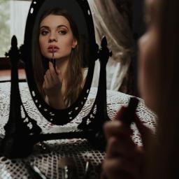 Dawid Mitoraj Fotografia Ślubna i Reportażowa - Młoda kobieta maluje usta szminką, patrząc w owalne lustro na stoliku z koronkowym obrusem, w tle okno z firanką.