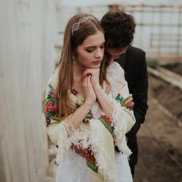 Dawid Mitoraj Fotografia Ślubna i Reportażowa - Panna młoda w koronkowej sukni i ludowym szalu z frędzlami, obejmowana przez pana młodego w czarnym garniturze na tle szklarni.