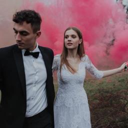 Dawid Mitoraj Fotografia Ślubna i Reportażowa - Para młoda w strojach ślubnych, panna młoda trzyma w dłoni czerwoną świecę dymną, w tle drzewa i różowy dym.