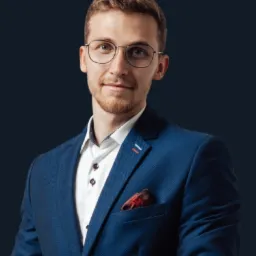 Portret młodego mężczyzny w okularach i niebieskiej marynarce, patrzącego wprost, na ciemnym tle. Widoczna koszula i ozdobna poszetka w kieszonce marynarki.