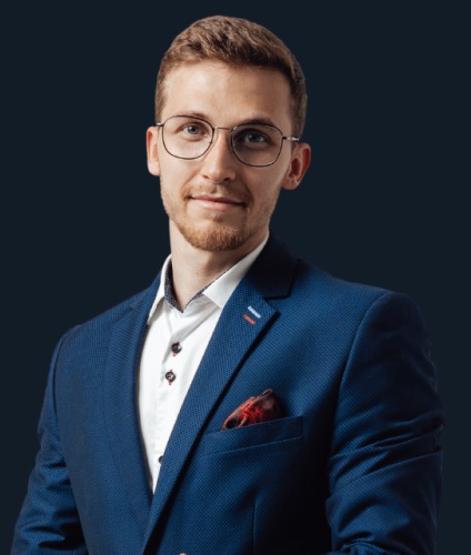 Portret młodego mężczyzny w okularach i niebieskiej marynarce, patrzącego wprost, na ciemnym tle. Widoczna koszula i ozdobna poszetka w kieszonce marynarki.