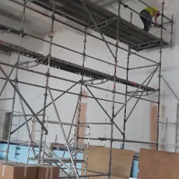 Pracownik w żółtej kamizelce na metalowym rusztowaniu wewnątrz budynku, widoczne przygotowane ściany, fragment basenu i kartony.