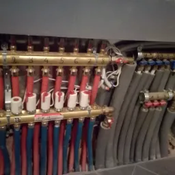 Rozdzielacz ogrzewania podłogowego z numerowanymi sekcjami, z widocznymi rurami w kolorach czerwonym i niebieskim, oraz izolowanymi rurami szarymi, zamontowany w surowym pomieszczeniu.