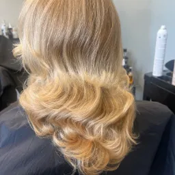 Tył głowy blondynki z pofalowanymi włosami po strzyżeniu z warstwami, widoczny czarny fartuch fryzjerski i fragment wyposażenia salonu.