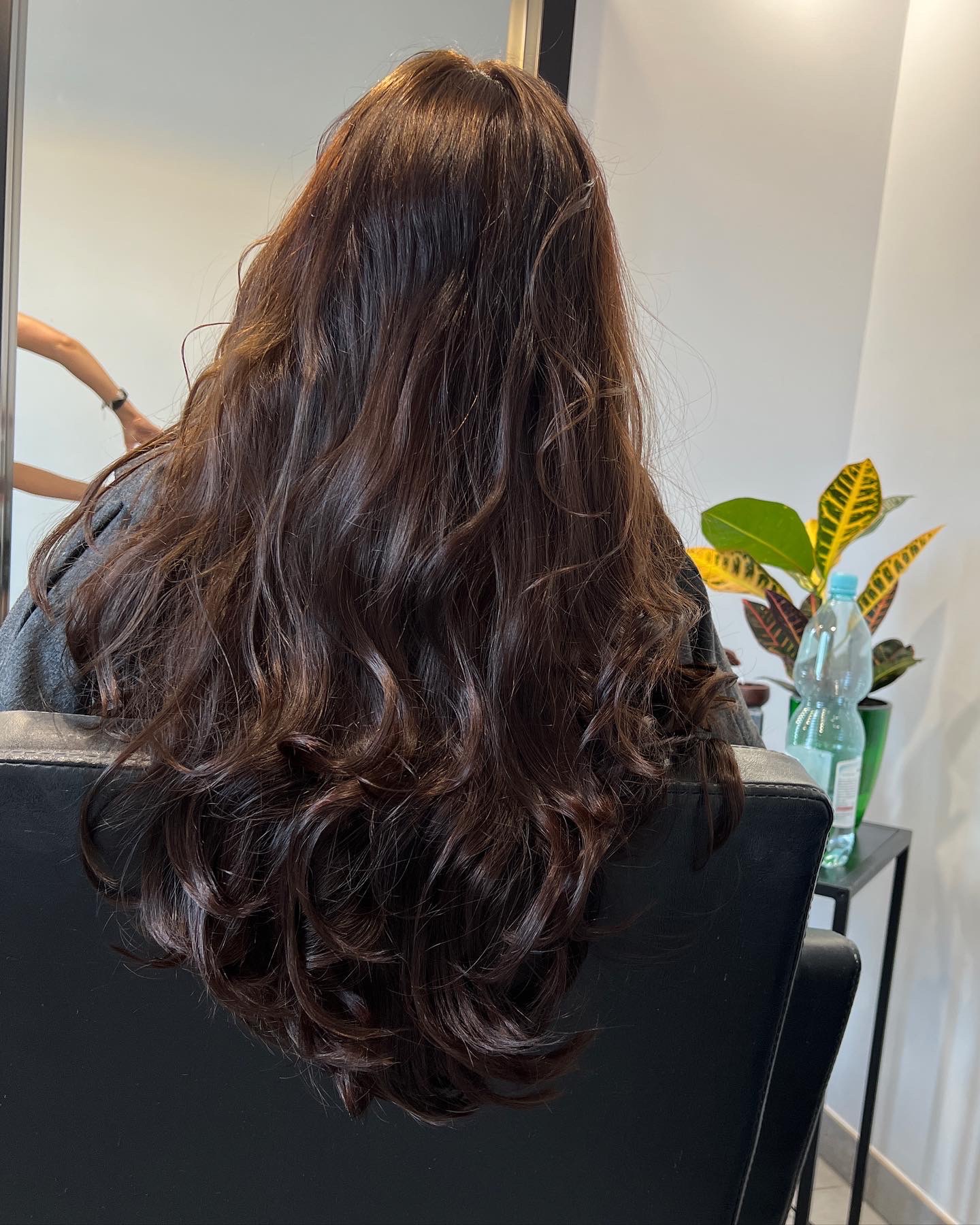 Długie, brązowe, falowane włosy na plecach klientki siedzącej w czarnym fotelu fryzjerskim, widok z tyłu, w tle lustro i roślina doniczkowa.