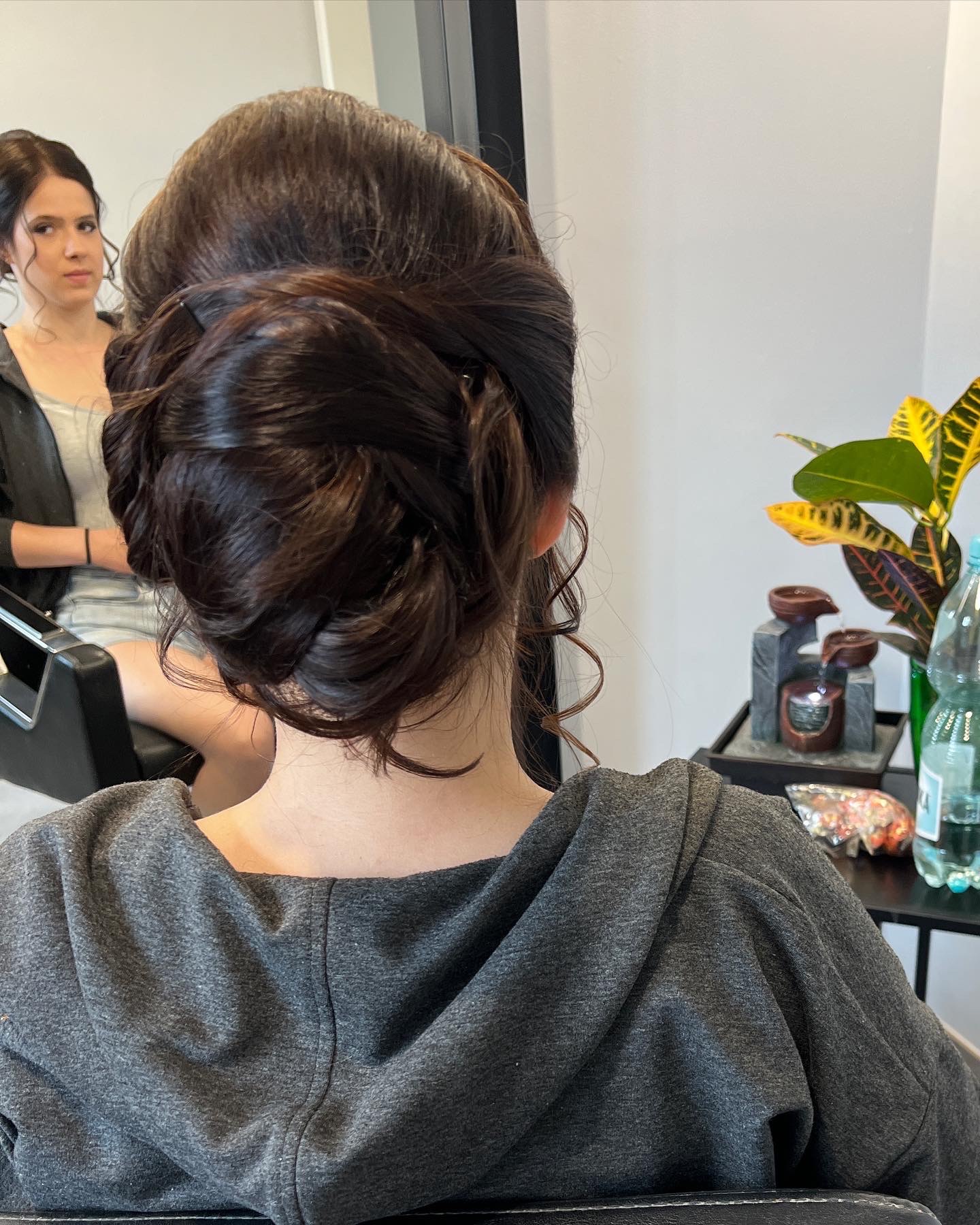 Upięcie włosów w kok z luźnymi pasmami, widok z tyłu na szyję klientki w szarej bluzie z kapturem, odbicie fryzjerki w lustrze w tle.