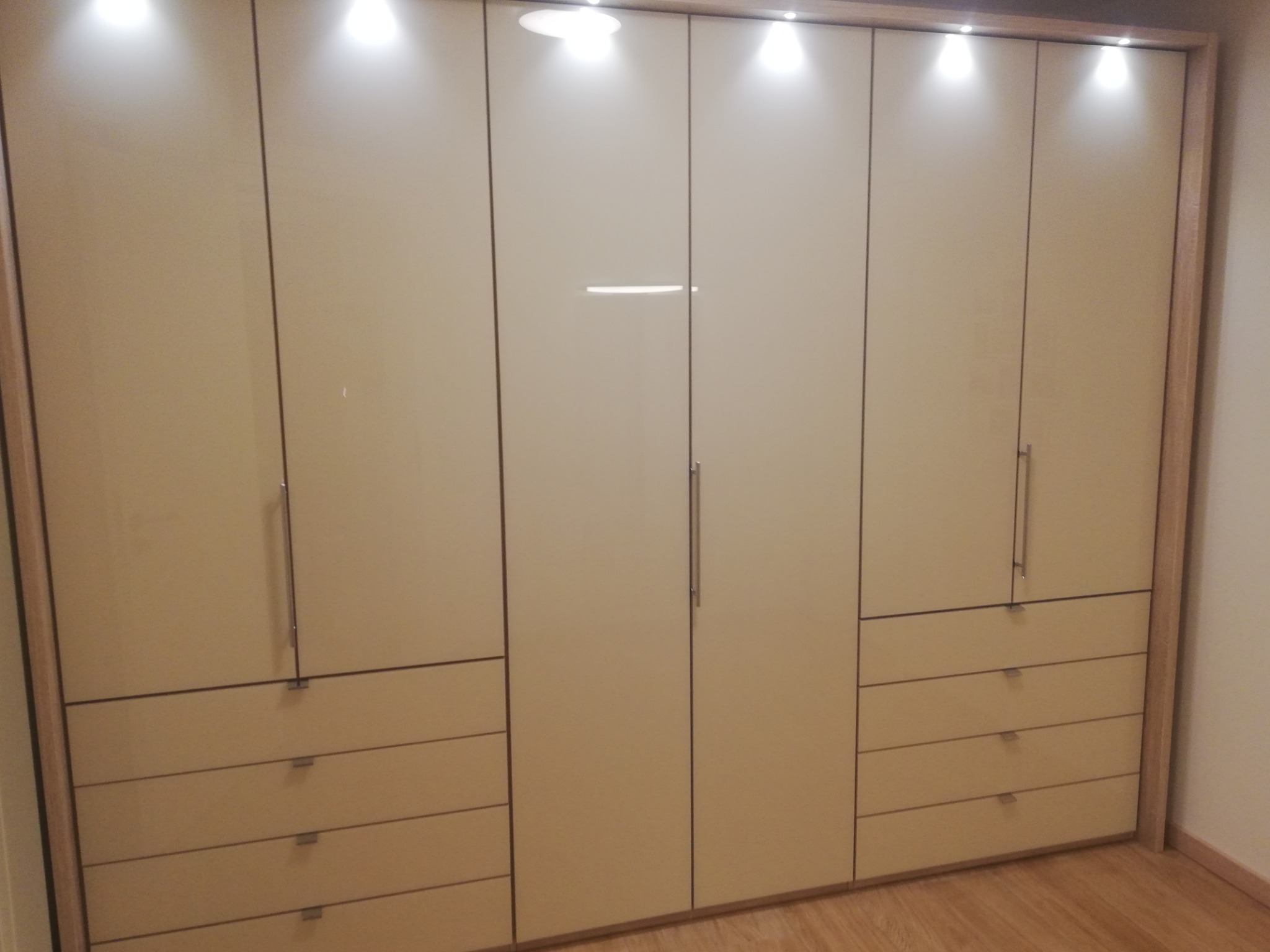 Beżowa szafa wnękowa z połyskującymi frontami, oświetleniem punktowym i czterema szufladami na dole, wykończona drewnianą ramą.